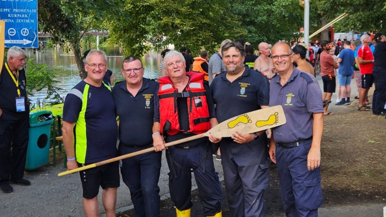 Fünf Männer in Uniform, zwei mit Rettungswesten, stehen am Fluss, lächeln und halten eine Paddel mit gelben Fußabdrücken.