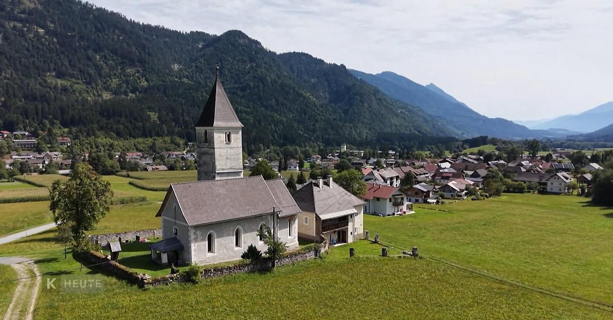 Eine kleine Dorfkirche mit einem Turm steht in einem üppigen grünen Feld mit Bergen im Hintergrund.