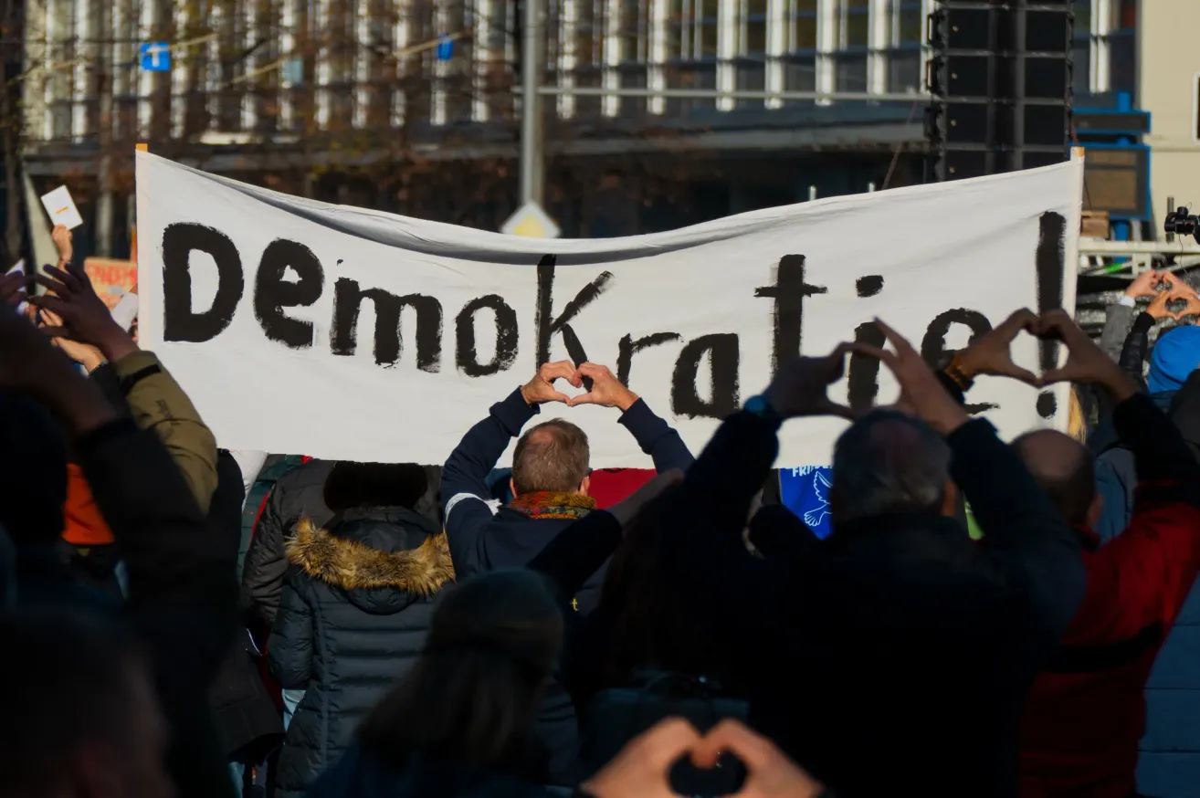 Demonstranten bilden mit ihren Händen ein Herz unter einem Banner, das Demokratie steht.