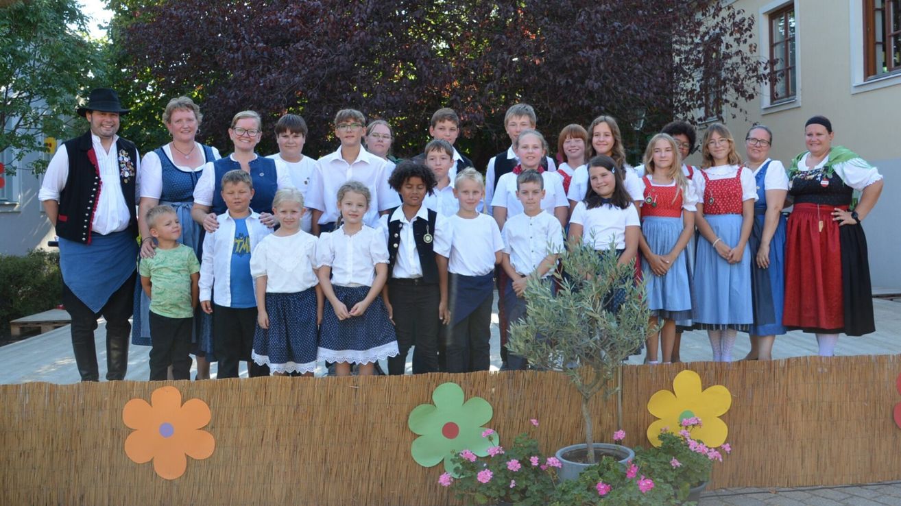 Eine Gruppe von Kindern und Erwachsenen in traditioneller Kleidung, die vor einem dekorativen Zaun mit Blumen und einem Baum stehen.
