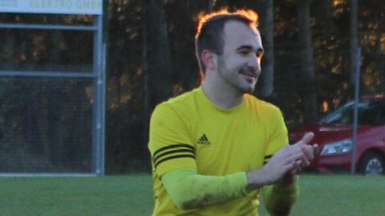 A man in a yellow Adidas shirt is smiling and gesturing with his hands on a grassy field.