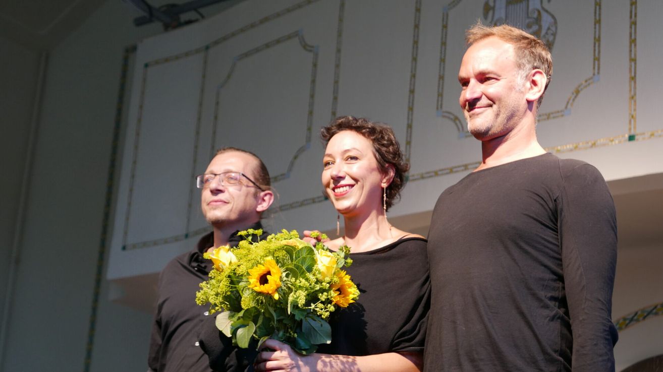 Three people stand together smiling, one holding a bouquet of sunflowers, another with glasses, and a third in a black shirt.