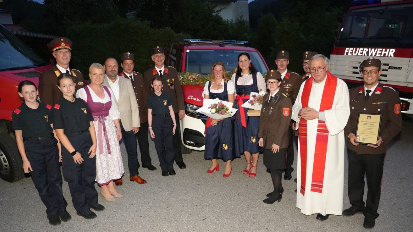 Eine Gruppe von Menschen, darunter uniformierte Beamte und Frauen in traditioneller Kleidung, steht vor einem roten Fahrzeug, möglicherweise einem Feuerwehrauto, mit Blumen und einem Priester im Hintergrund.