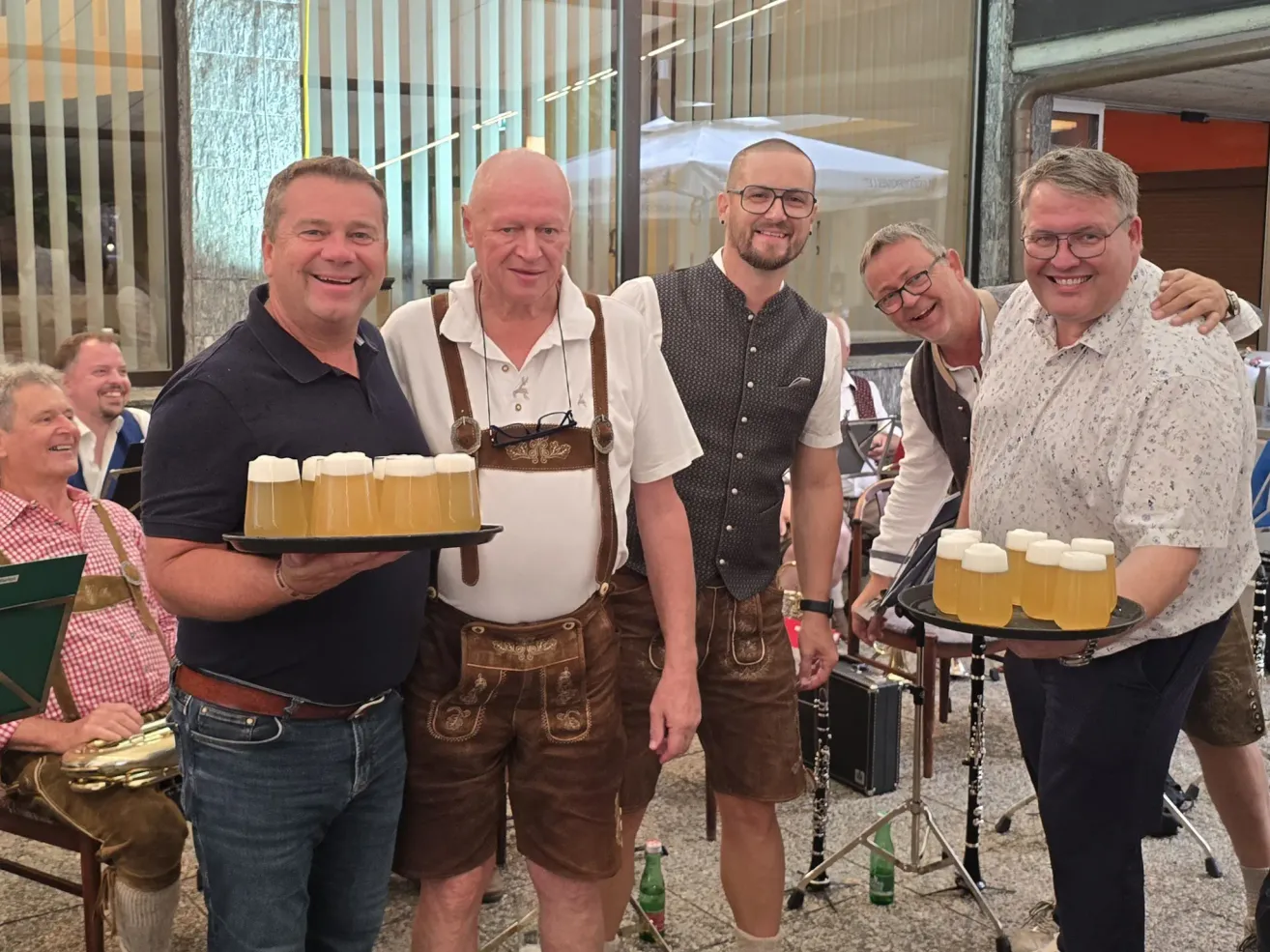 Fünf Männer in traditioneller bayerischer Kleidung lächeln und halten Tabletts mit Biergläsern. Der Mann auf der linken Seite trägt ein schwarzes Shirt und Jeans. Der Mann neben ihm trägt ein weißes Shirt, braune Träger und braune Shorts. Der Mann in der Mitte trägt einen schwarzen Weste und Brille. Der Mann auf der rechten Seite trägt ein weißes Shirt und Brille.