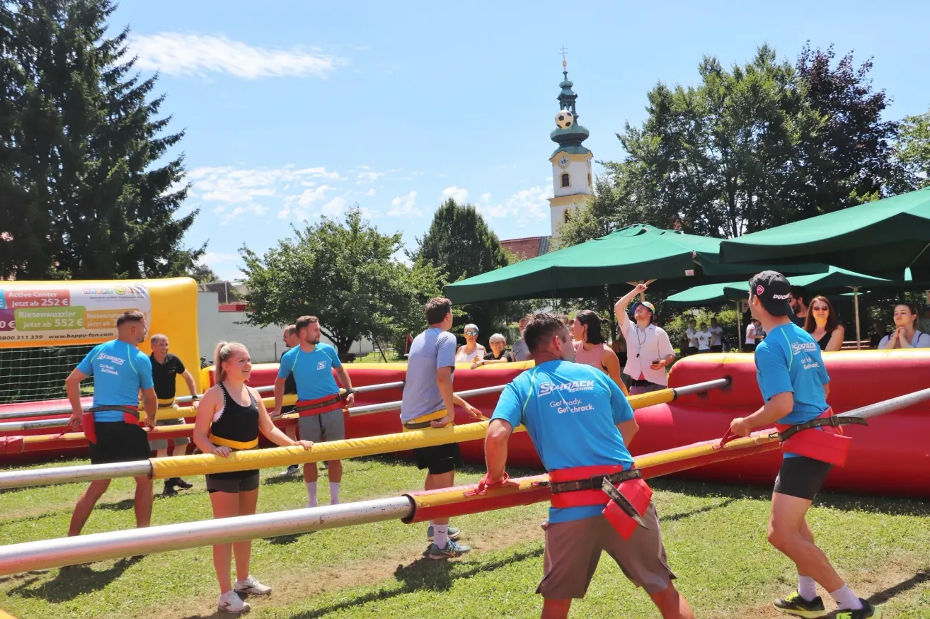 Eine Gruppe von Menschen nimmt an einer Teambuilding-Aktivität im Freien teil, wobei sie gelbe und rote Stangen halten, mit einem Kirchturm und Bäumen im Hintergrund.