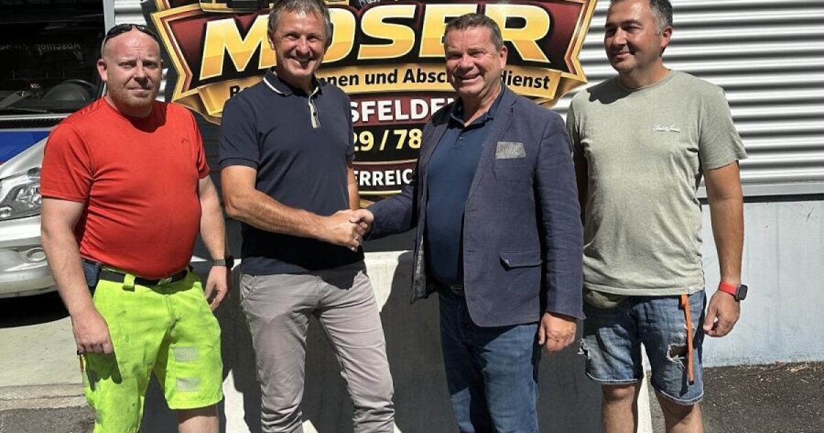 Vier Männer stehen vor einer Wand mit einem Schild, auf dem 'MOSER' steht. Der Mann auf der linken Seite trägt ein rotes Shirt und neongrüne Shorts. Der Mann in der Mitte schüttelt dem Mann auf der rechten Seite die Hand, der eine hellblaue Jacke trägt.