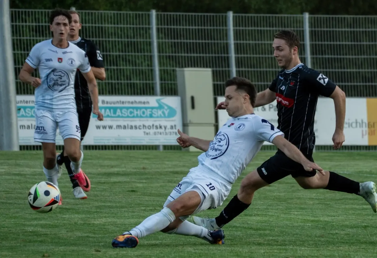 Two soccer players in white and black uniforms are in a match on a grassy field. One player is sliding on the ground while the other is trying to defend. Behind them is a fence with a signboard.