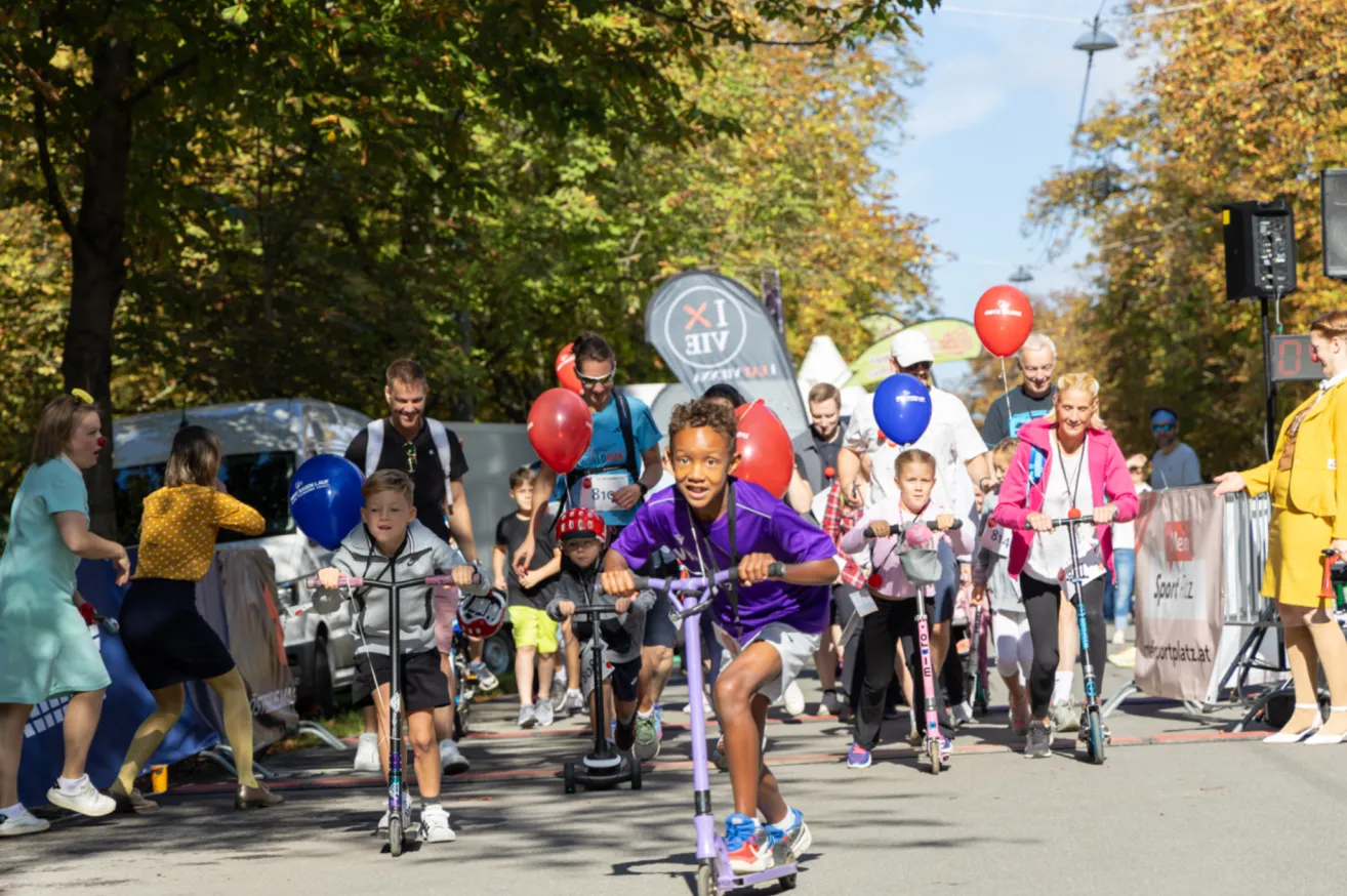 A group of children and adults ride scooters in a parade, with colorful balloons and a banner in the background.