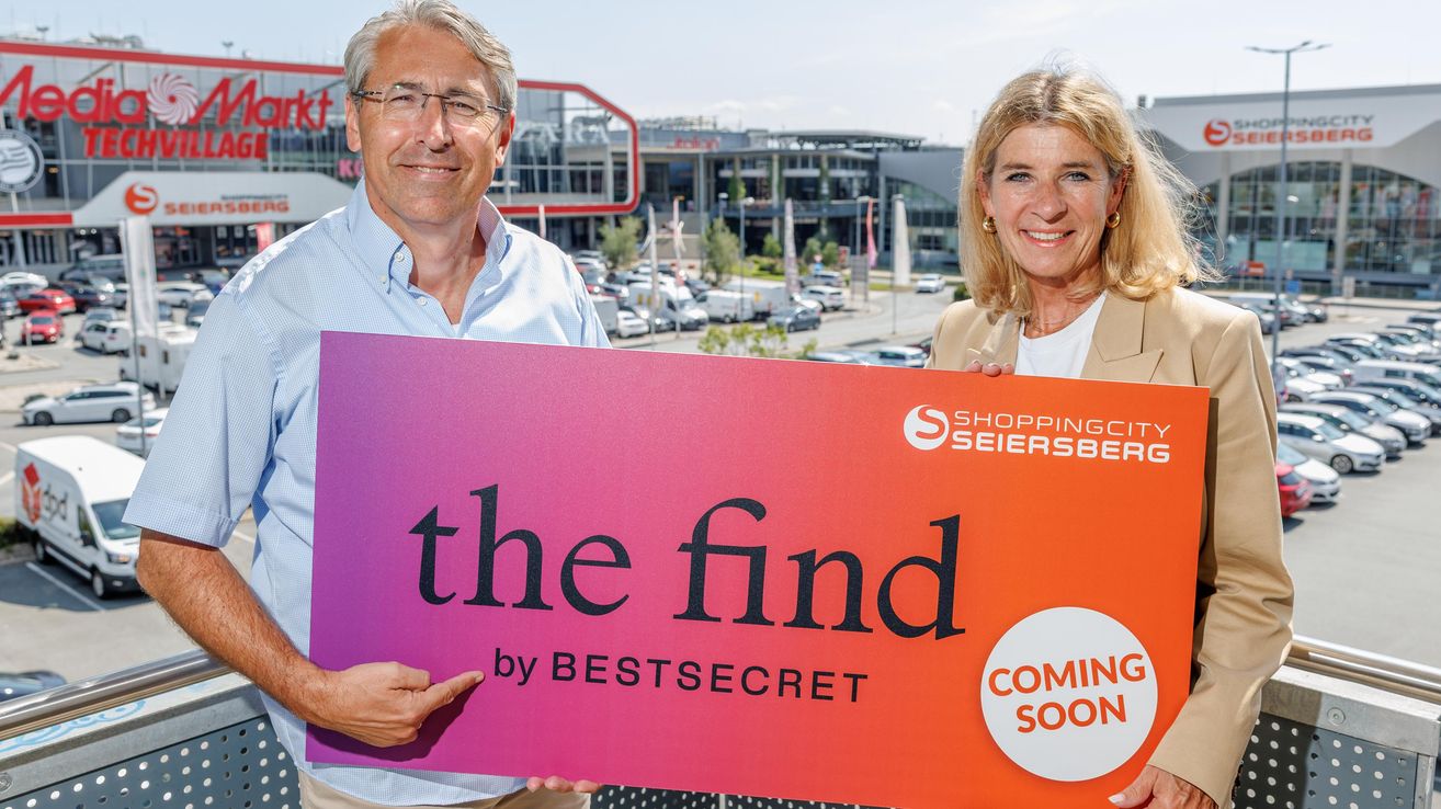 A man and a woman are holding a large sign that reads 'the find by BESTSECRET' in front of a shopping center.