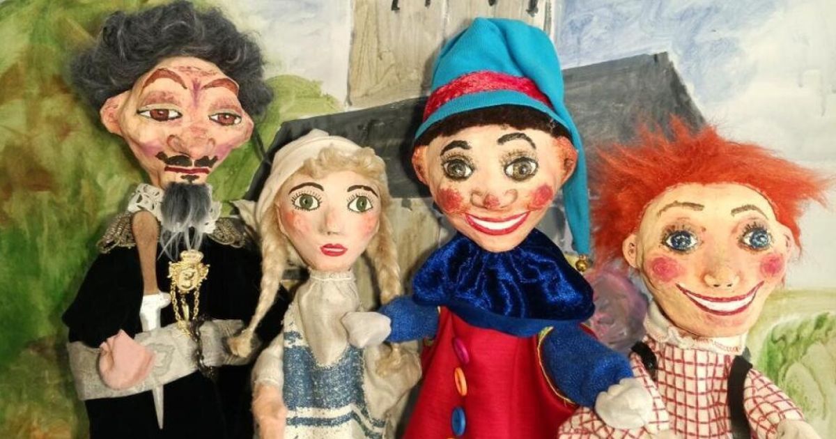 Vier handgefertigte Puppen mit unterschiedlichen Gesichtsausdrücken und Kostümen stehen zusammen. Die linkeste Puppe trägt einen schwarzen Umhang und eine goldene Kette. Daneben eine blonde Puppe in einem blauen Kleid. Die mittlere Puppe trägt einen blauen Hut und ein rotes Kostüm. Die rechte Puppe hat rote Haare und ein weiß-rotes kariertes Hemd.
