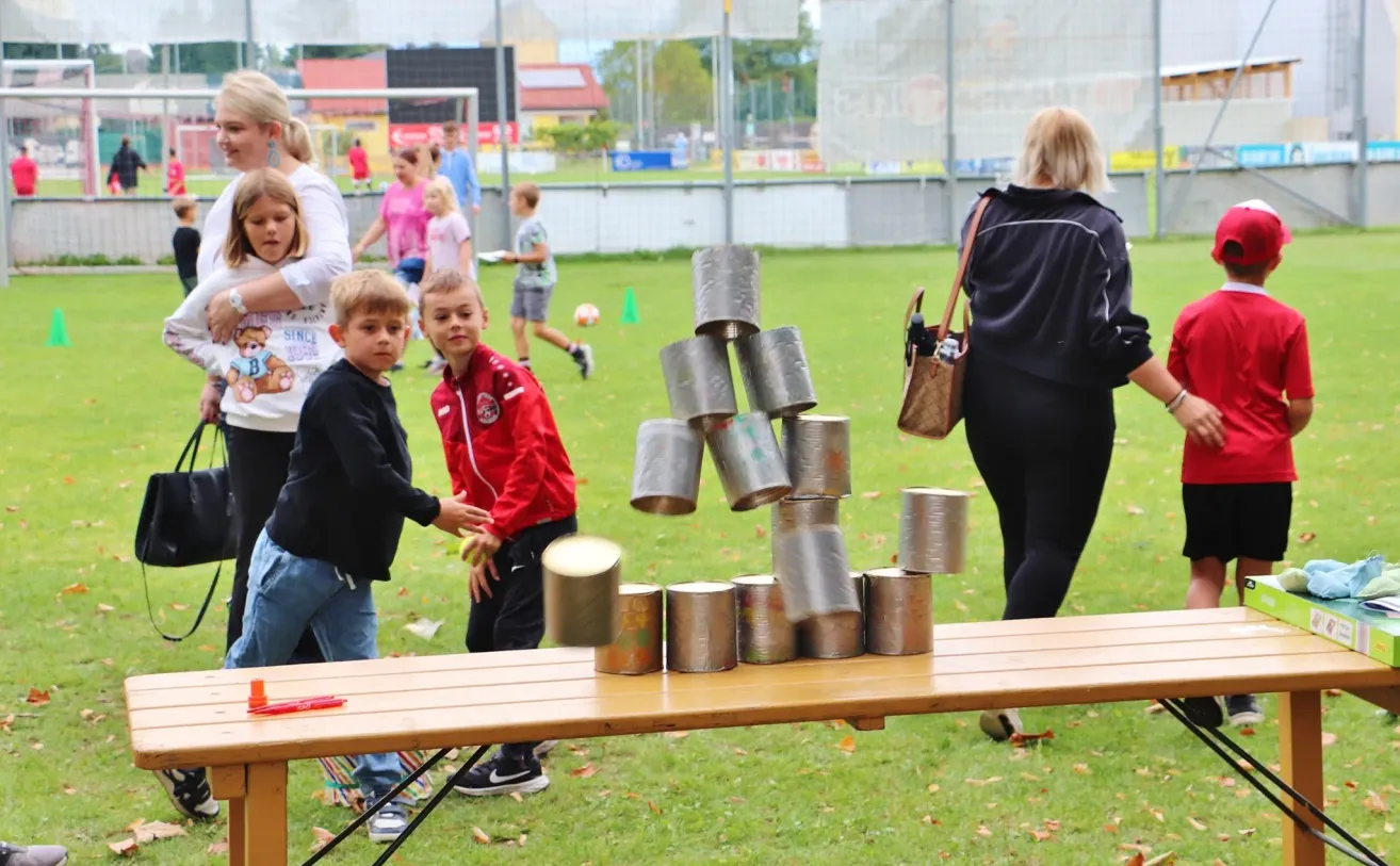 Children are playing with a stack of metal cans on a table in a park. A woman is walking nearby.
