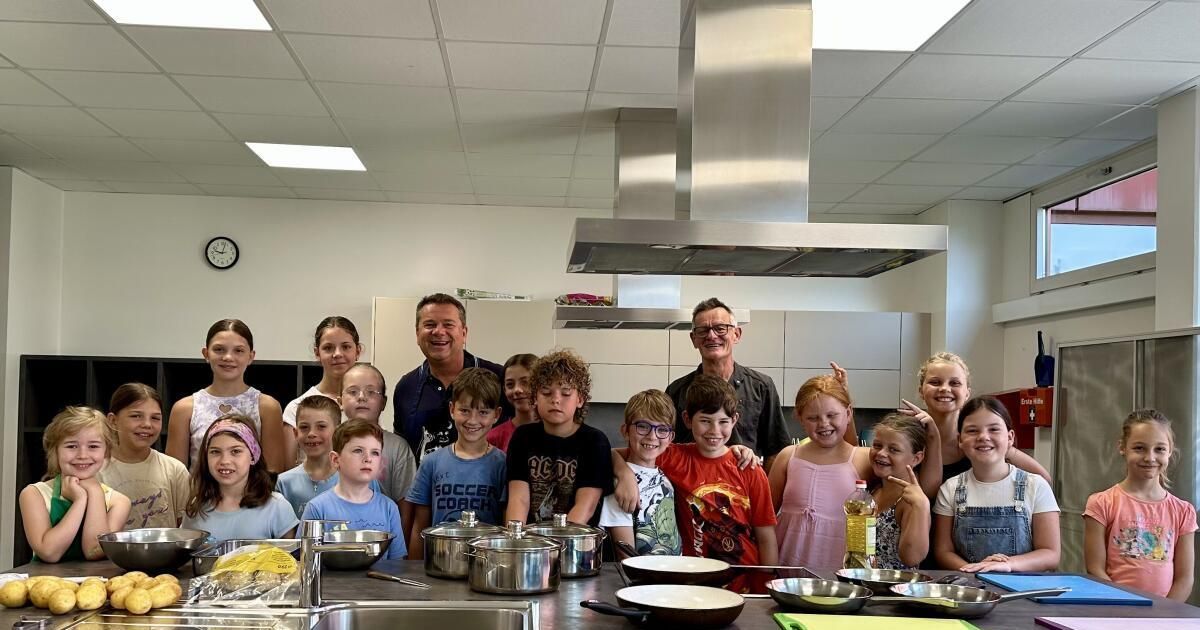 Eine Gruppe von Kindern und zwei Erwachsenen posiert für ein Foto in einer Küche mit Edelstahltöpfen und -pfannen auf dem Tresen.