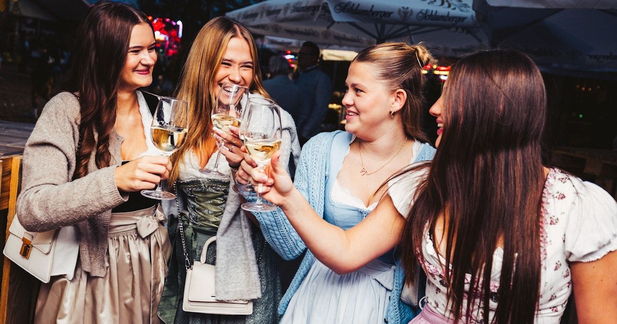 Vier Frauen in traditioneller deutscher Kleidung stoßen mit ihren Weinläsern bei einer Veranstaltung im Freien an, lächeln und lachen.
