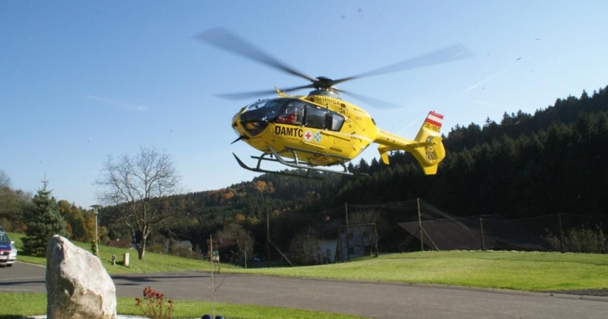 A yellow helicopter with DAMTC and a red cross on the side is flying above a grassy field with a house and trees in the background.