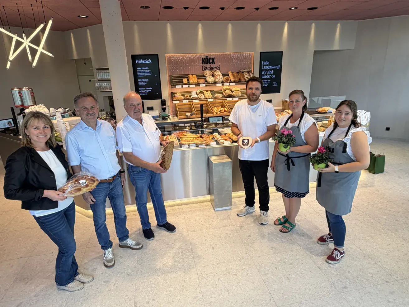 Fünf Personen stehen lächelnd vor einer Bäckereitheke und halten Brot. Die Bäckerei hat ein modernes Design mit Regalen voller Brot und Gebäck.