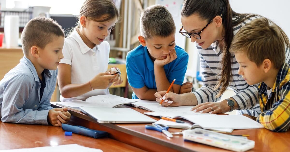 Eine Lehrerin hilft einer Gruppe von Schülern im Klassenzimmer, wobei ein Mädchen ein kleines Objekt hält, ein Junge schreibt und ein anderer Junge nach unten schaut.