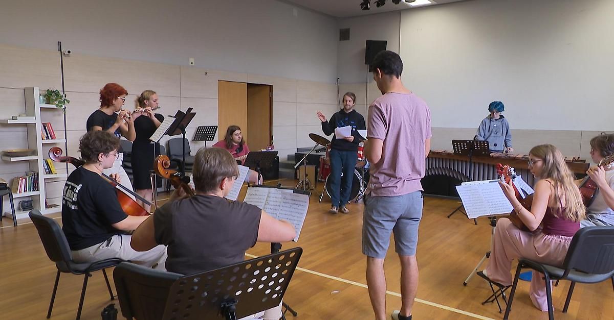 Eine Gruppe von Musikern übt in einem Raum mit Holzböden. Einige sitzen und spielen Instrumente, während andere stehen und einem Mann zuhören.