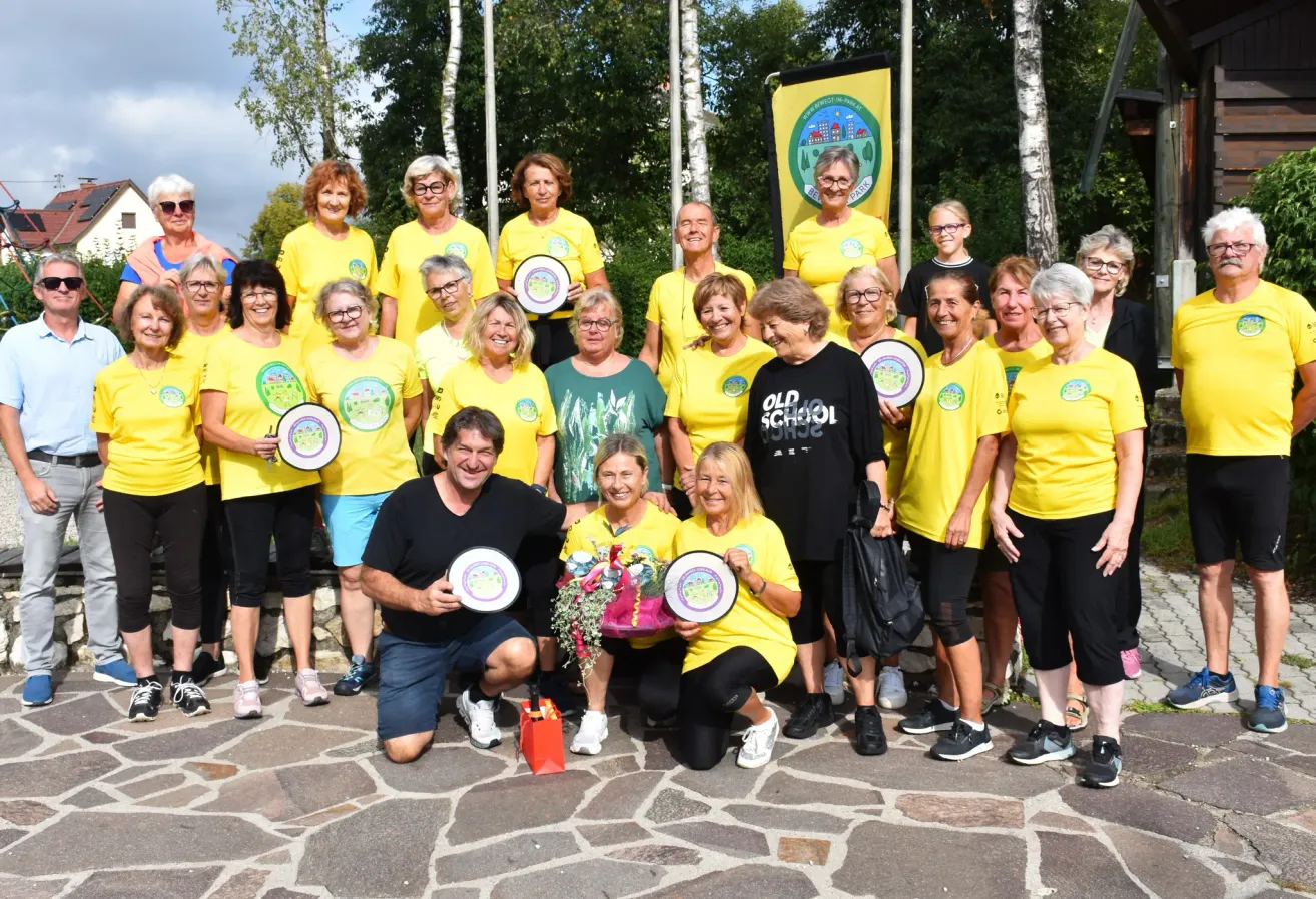 Eine Gruppe von Menschen in gelben T-Shirts posiert für ein Foto, einige halten Teller und Blumen. Ein Mann in einem schwarzen Shirt kniet vor ihnen.