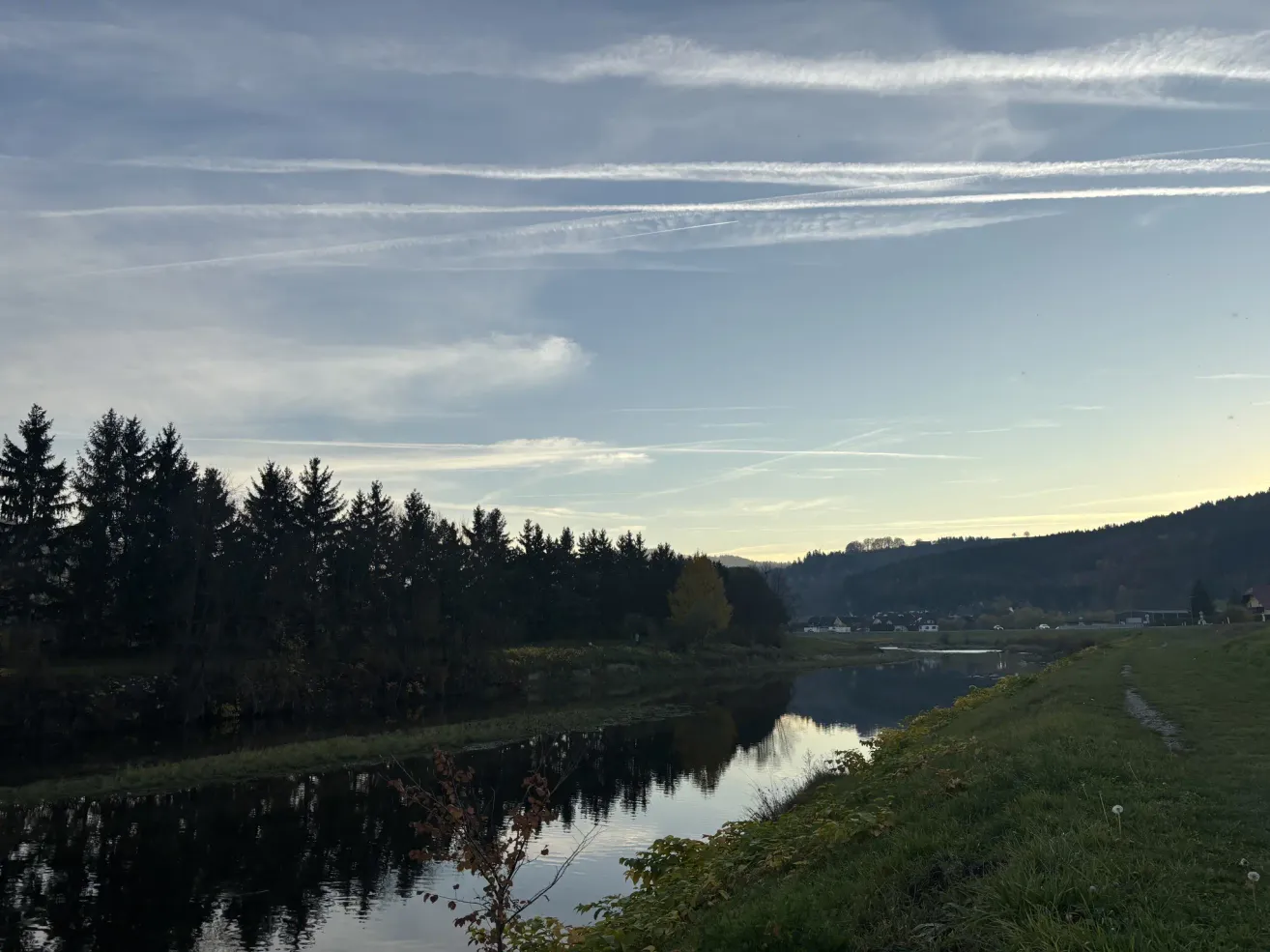Eine friedliche Landschaft mit einem ruhigen Fluss, dichtem Wald und einem klaren blauen Himmel mit flaumigen Wolken.