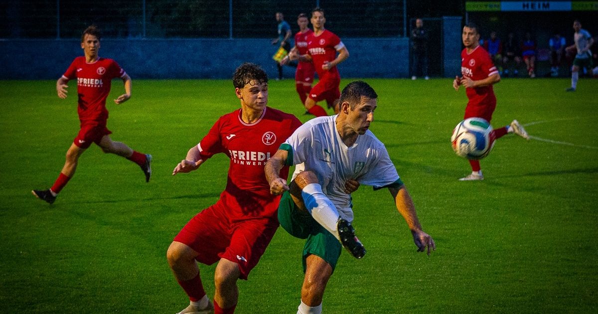 Zwei Fußballspieler in roten und grünen Trikots sind auf dem Feld in einen körperlichen Zweikampf verwickelt. Der Spieler in Rot drängt den Spieler in Grün, der versucht, das Gleichgewicht zu halten. Andere Spieler sind im Hintergrund, und ein Fußball ist in der Luft.