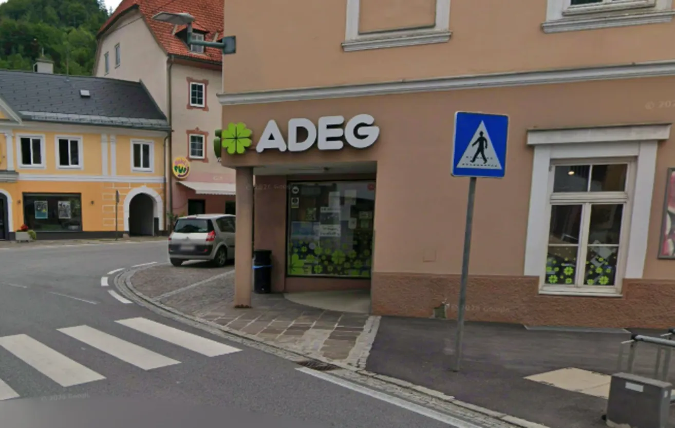 Ein Laden mit einem Schild, das ADEG liest, ein Fußgängerübergangsschild und ein geparktes Auto im Hintergrund.