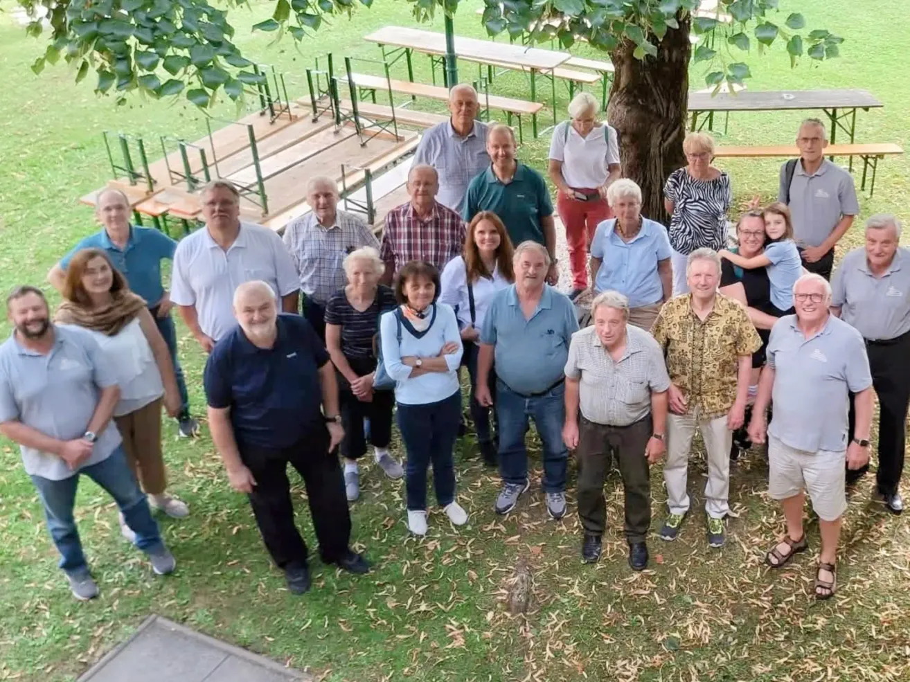 Eine Gruppe von Erwachsenen und Kindern, die in einem Park stehen, mit einem Baum in der Mitte und Picknicktischen im Hintergrund.