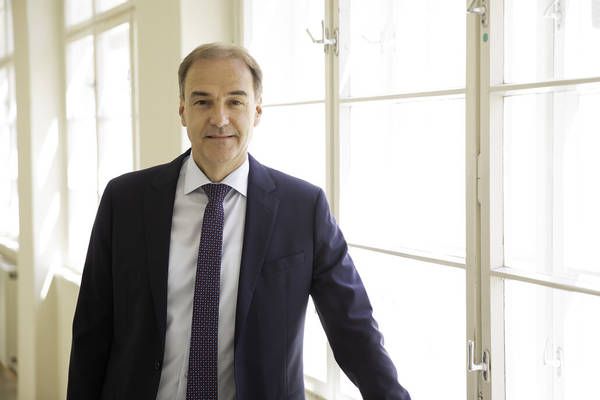 A man in a business suit stands confidently in a room with large windows, smiling at the camera.