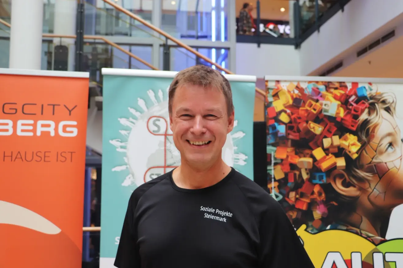A man wearing a black shirt with the words 'Soziale Projekte Steiermark' smiles in front of colorful banners.