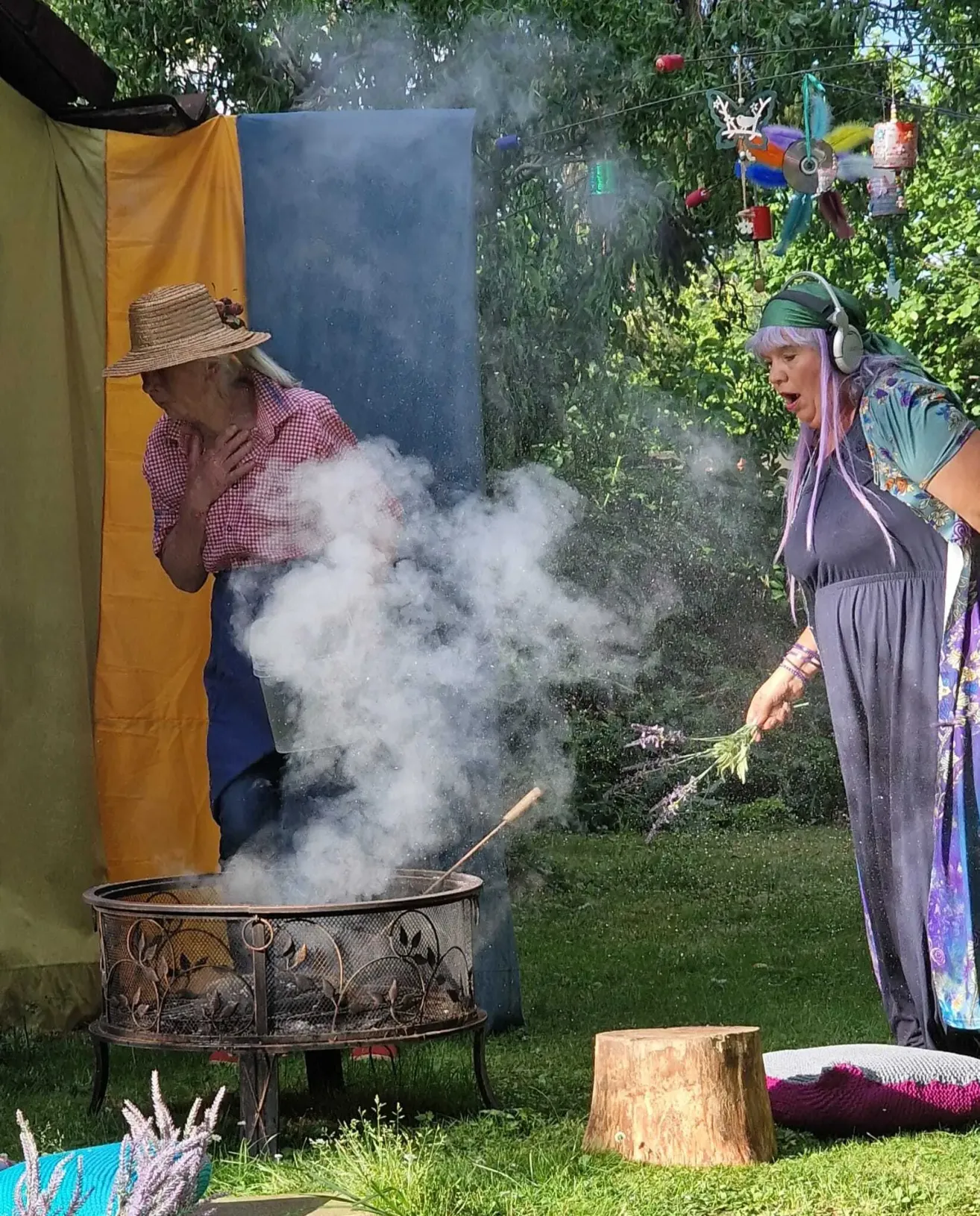 Two women stand in a garden, one blowing smoke from a stick while the other holds lavender and sings.