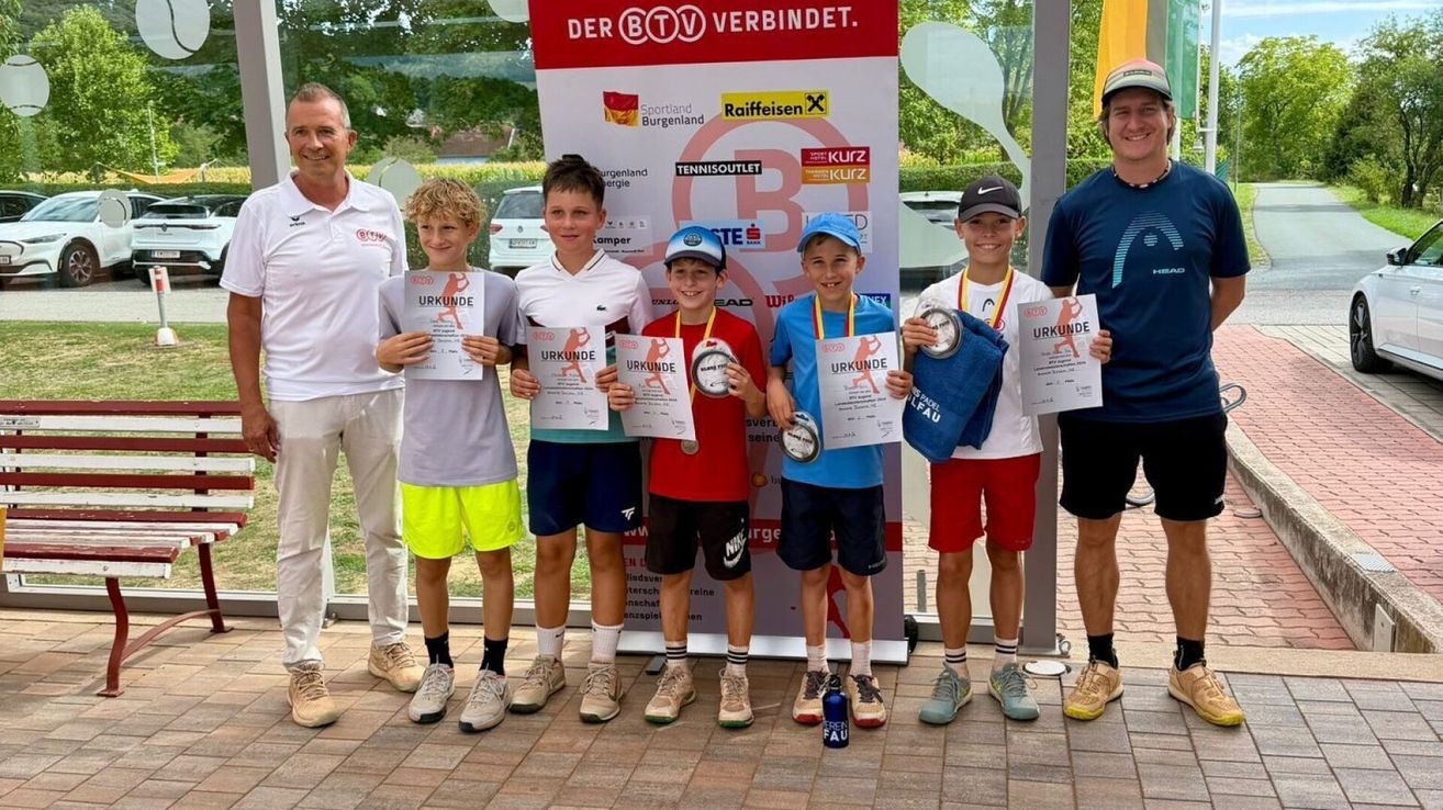 Fünf junge Jungen in Sportkleidung stehen vor einem Banner, halten Urkunden und Medaillen in den Händen. Sie lächeln und posieren für ein Foto. Dahinter stehen zwei Erwachsene, einer von ihnen trägt einen Hut. Im Hintergrund befindet sich eine Rasenfläche und ein Auto.