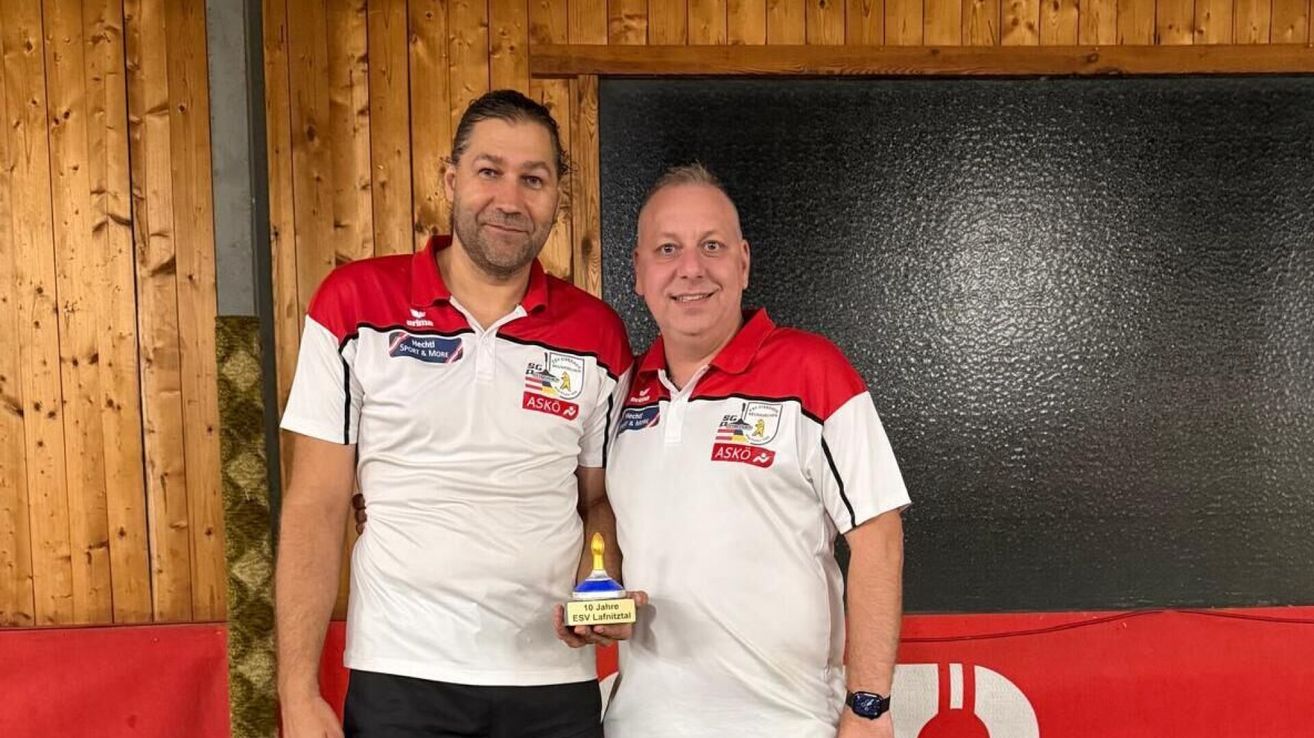 Zwei Männer stehen nebeneinander, lächeln und halten eine Trophäe mit der Aufschrift '10 Jahre ESV Lahntal'. Sie tragen passende rote und weiße Polo-Shirts mit Logos und Text auf der Brust. Dahinter befindet sich eine Holzwand und eine schwarze Oberfläche.