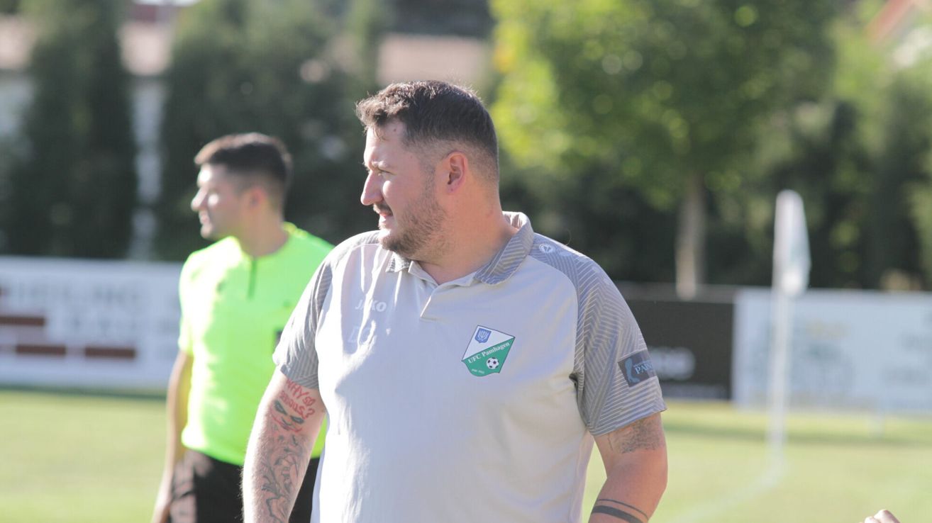 Ein Mann mit Tattoos an den Armen trägt ein weißes und graues Shirt mit einem Fußballlogo auf der linken Brust auf einem Fußballfeld.