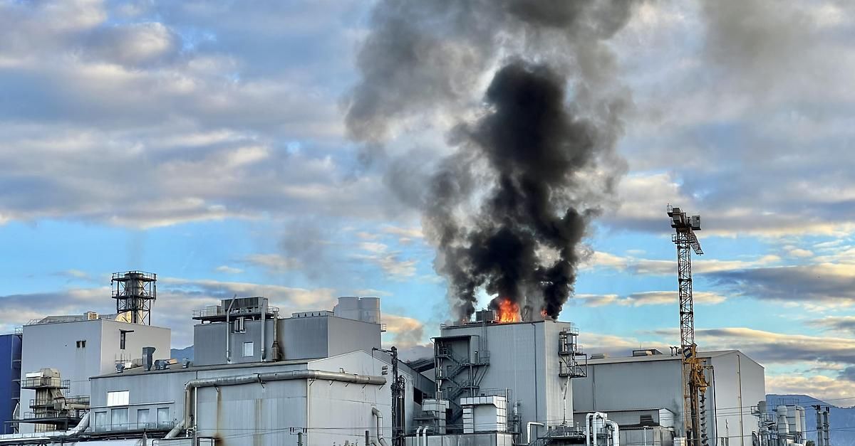 A factory with a fire on its roof, emitting thick black smoke against a blue sky with scattered clouds.