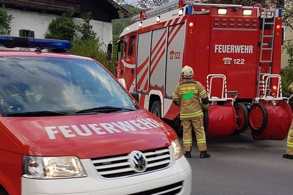 A red fire truck with the word 'Feuerwehr' on it is parked on the side of the road. A firefighter is standing next to it, and a police car is parked nearby.