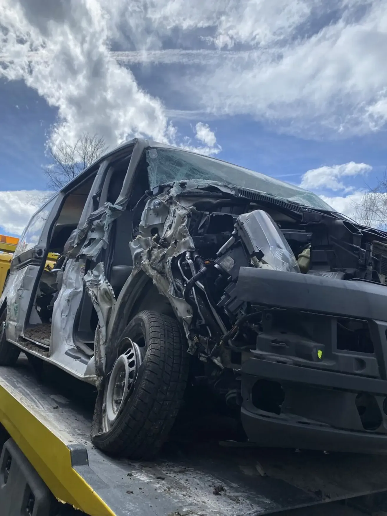 A severely damaged vehicle is being towed on a flatbed truck. The front is completely crushed, with the hood and bumper detached. The windshield is shattered, and the interior is exposed.