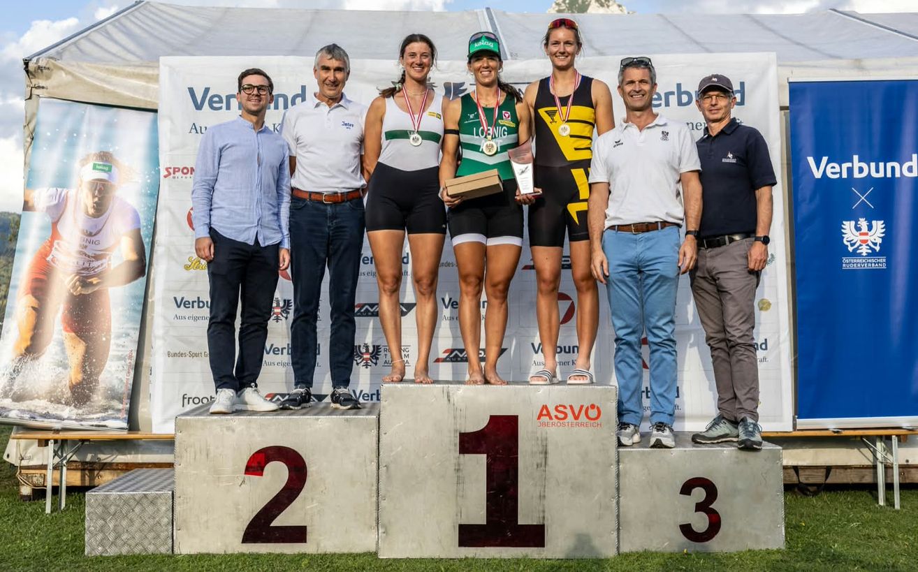 Drei Frauen stehen auf einem Podest mit Medaillen um den Hals, während zwei Männer hinter ihnen stehen. Die Frau in der Mitte hält eine Trophäe. Das Podest hat die Zahlen 1, 2 und 3 darauf.