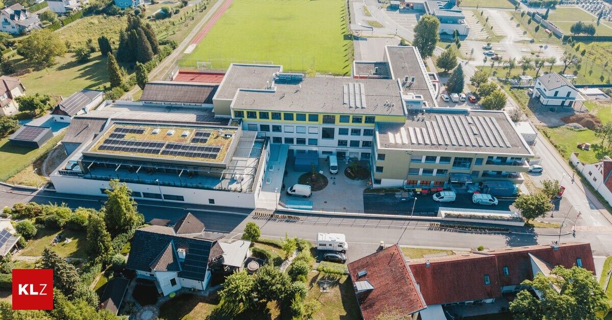 Ein Luftbild eines modernen Gebäudekomplexes mit Solarmodulen auf dem Dach, umgeben von Grünflächen und einem Parkplatz mit mehreren Fahrzeugen.