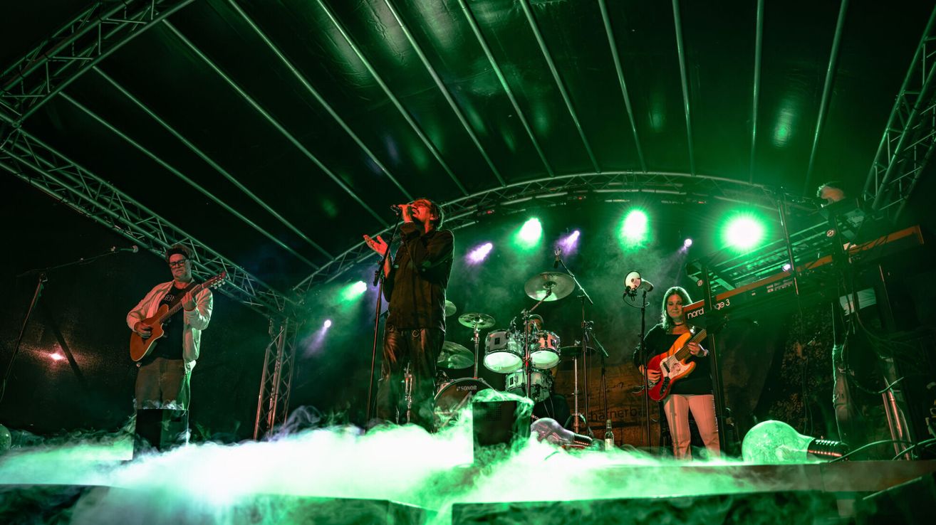Eine Rockband tritt auf der Bühne mit grüner Beleuchtung, Rauch und einem Mikrofon auf. Der Leadsänger steht in der Mitte, während andere Schlagzeug und Gitarre spielen.