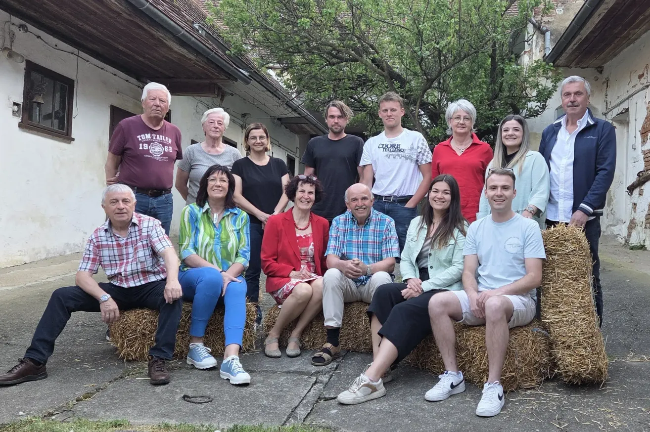 Eine Gruppe von Menschen, sowohl Männer als auch Frauen, sitzen auf Heuballen vor einem Gebäude. Sie alle lächeln und scheinen für ein Foto zu posieren.