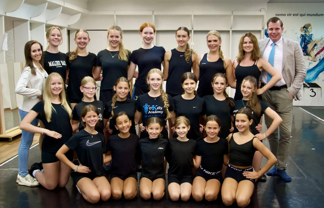 Eine Gruppe junger Mädchen, wahrscheinlich ein Sportteam, gekleidet in schwarzen Uniformen, posiert für ein Foto im Studio. Einige knien, andere stehen und alle scheinen zu lächeln.
