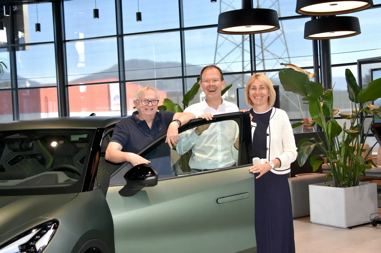Drei Personen stehen neben einem Auto in einer Ausstellung. Der Mann links trägt eine Brille und eine Uhr. Der Mann in der Mitte trägt ein weißes Hemd und die Frau rechts trägt eine weiße Strickjacke.