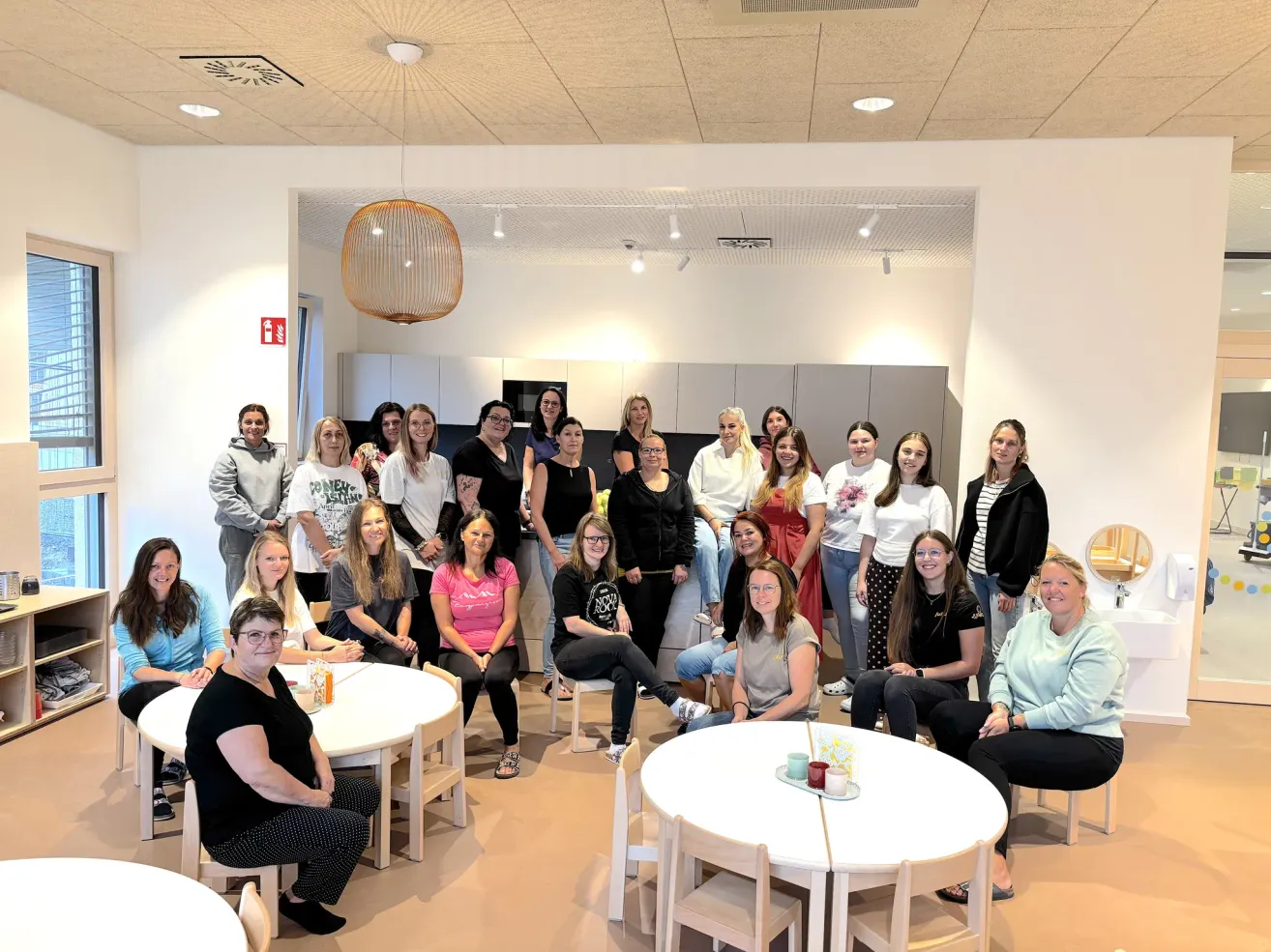 Eine Gruppe von Frauen versammelt sich in einem Raum, einige sitzen auf Stühlen und andere stehen, mit einem Tisch in der Mitte. Sie sind lässig gekleidet und scheinen für ein Foto zu posieren. Der Raum hat weiße Wände und eine Decke mit Lichtern.