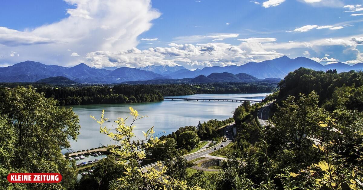 Ein ruhiger See mit einer Brücke, umgeben von Bergen und üppigem Grün, unter einem blauen Himmel mit verstreuten Wolken.