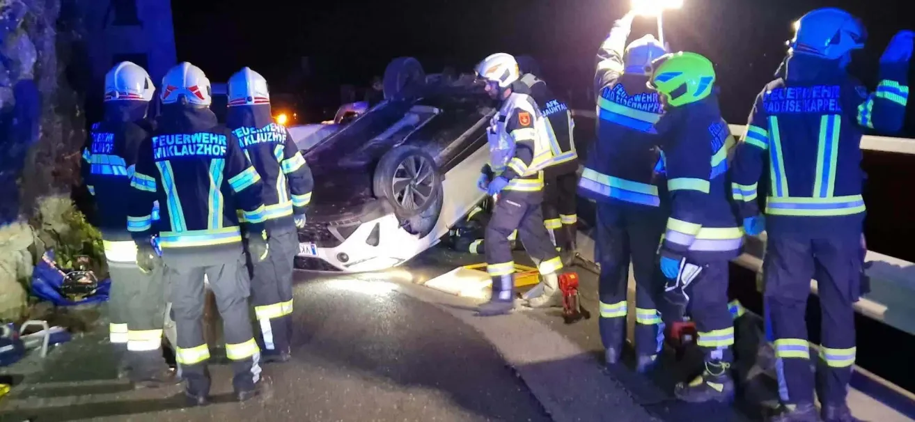 Einsatzkräfte kümmern sich um ein umgekipptes Auto auf einer Straße bei Nacht. Ein Helfer trägt einen weißen Helm und blaue Handschuhe.