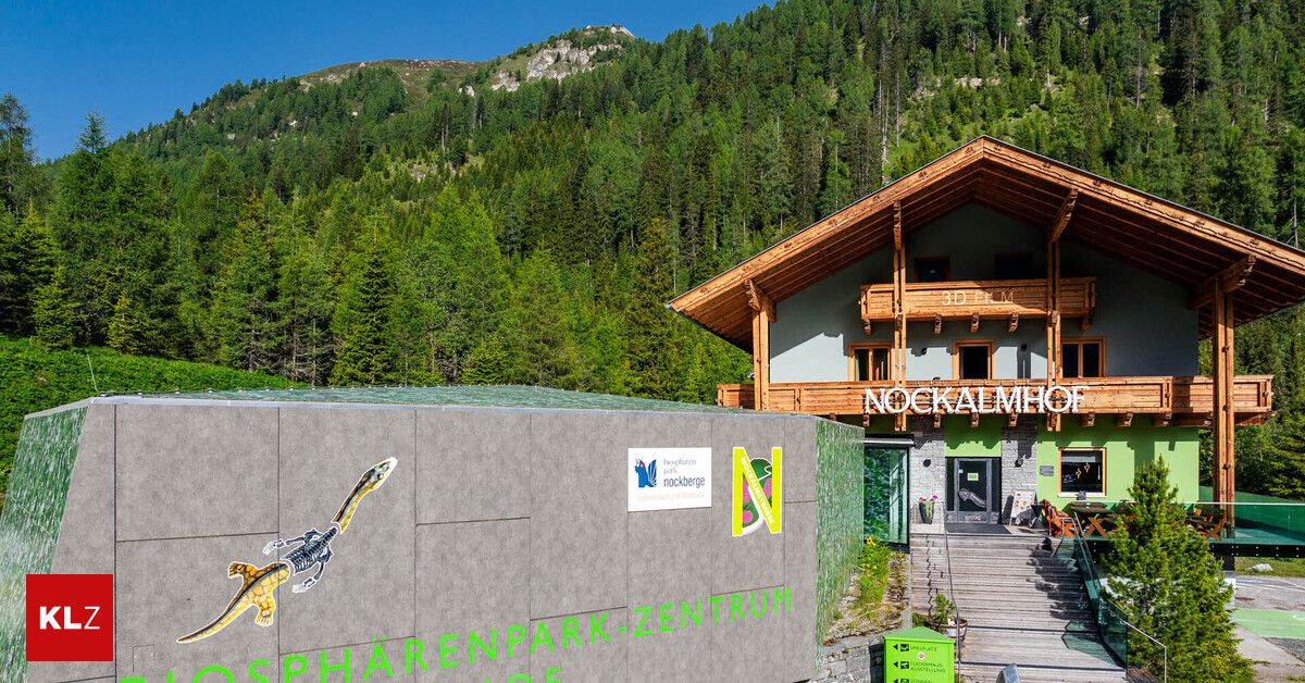 Ein Gebäude mit einem hölzernen Balkon und einem Schild mit der Aufschrift 'NOCKALMHOCH' steht vor einem Wald. Das Gebäude hat eine grüne Wand mit dem Text 'SPHARENPARK-ZENTRUM' und einer Eidechsenzeichnung.