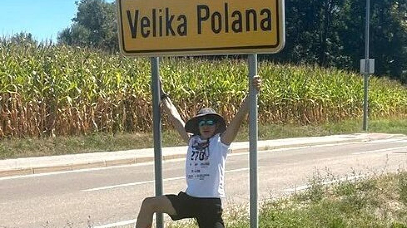 A person wearing a hat and sunglasses poses for a photo at a road sign for Velika Polana.