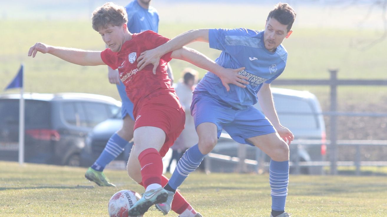 Two soccer players in red and blue uniforms are engaged in a physical duel on a grassy field. One player is about to kick the ball while the other defends.