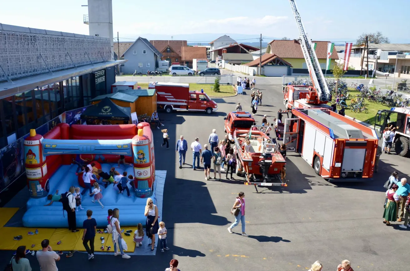 A community event with a fire truck, inflatable bounce house, and people walking around. Buildings and parked cars are in the background.