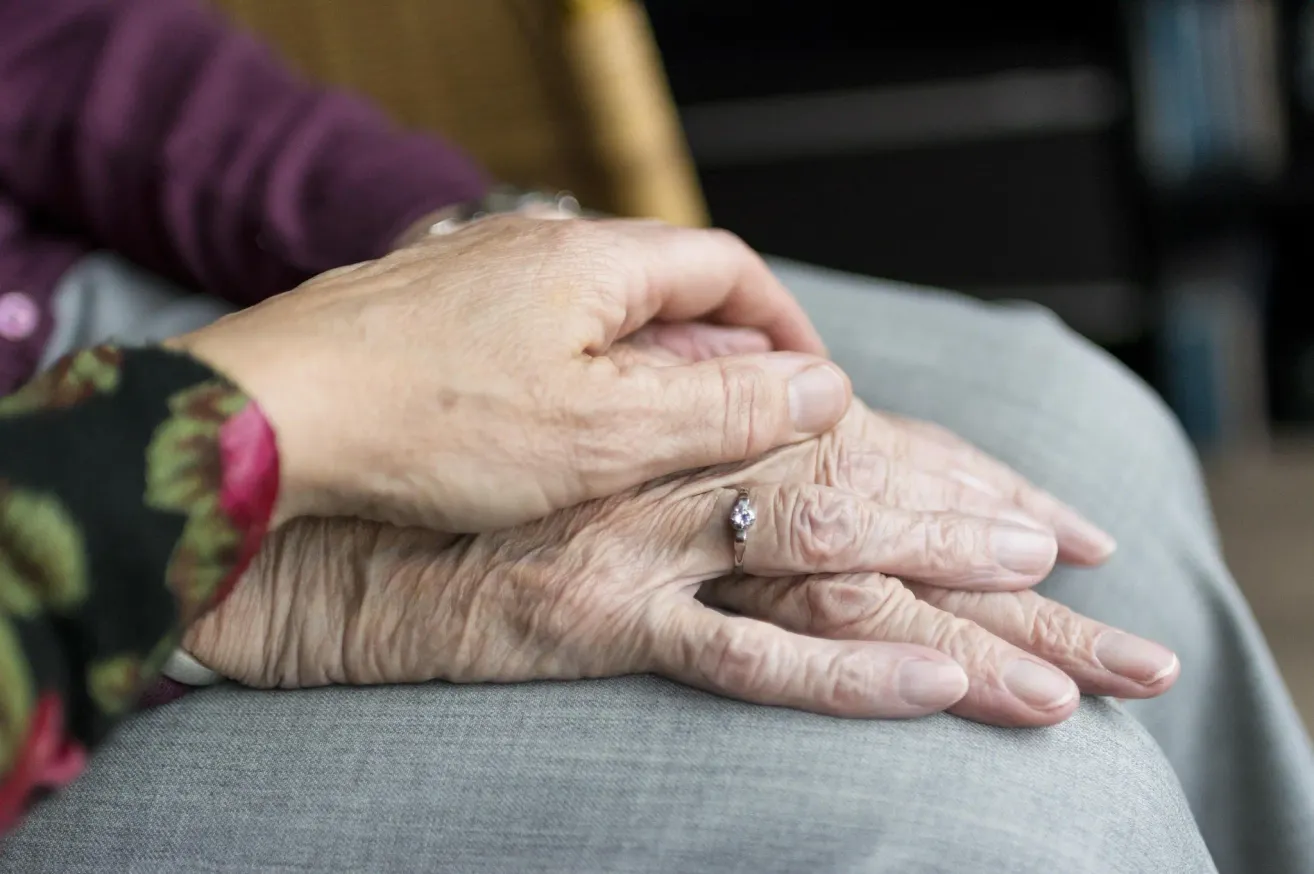 An elderly person's hands are clasped together, with a ring on the middle finger of the right hand.
