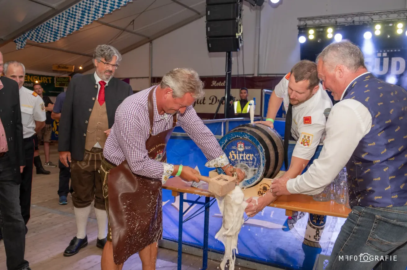 Ein Mann in einer Lederschürze klopft mit einem Holzhammer auf einen großen Fass, während zwei andere ihm helfen. Sie befinden sich auf einem Festival mit einem blauen Hintergrund und einem sichtbaren Lautsprecher.